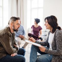 Senior counselor with clipboard talking to a man during group therapy.
